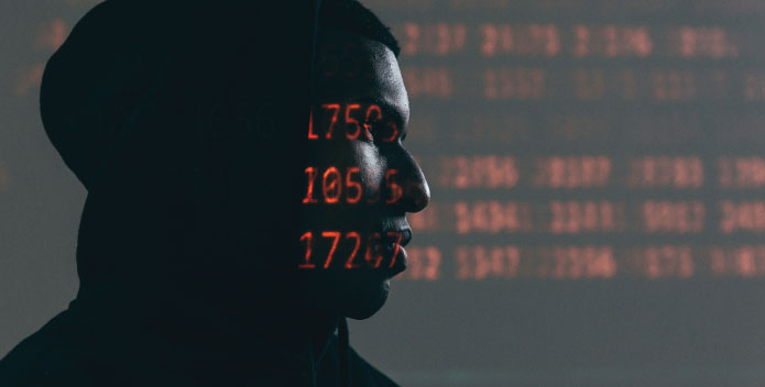 Silhoutte of a suspicious looking man with code projected on to his face to illustrate the need for Cybercrime Defence